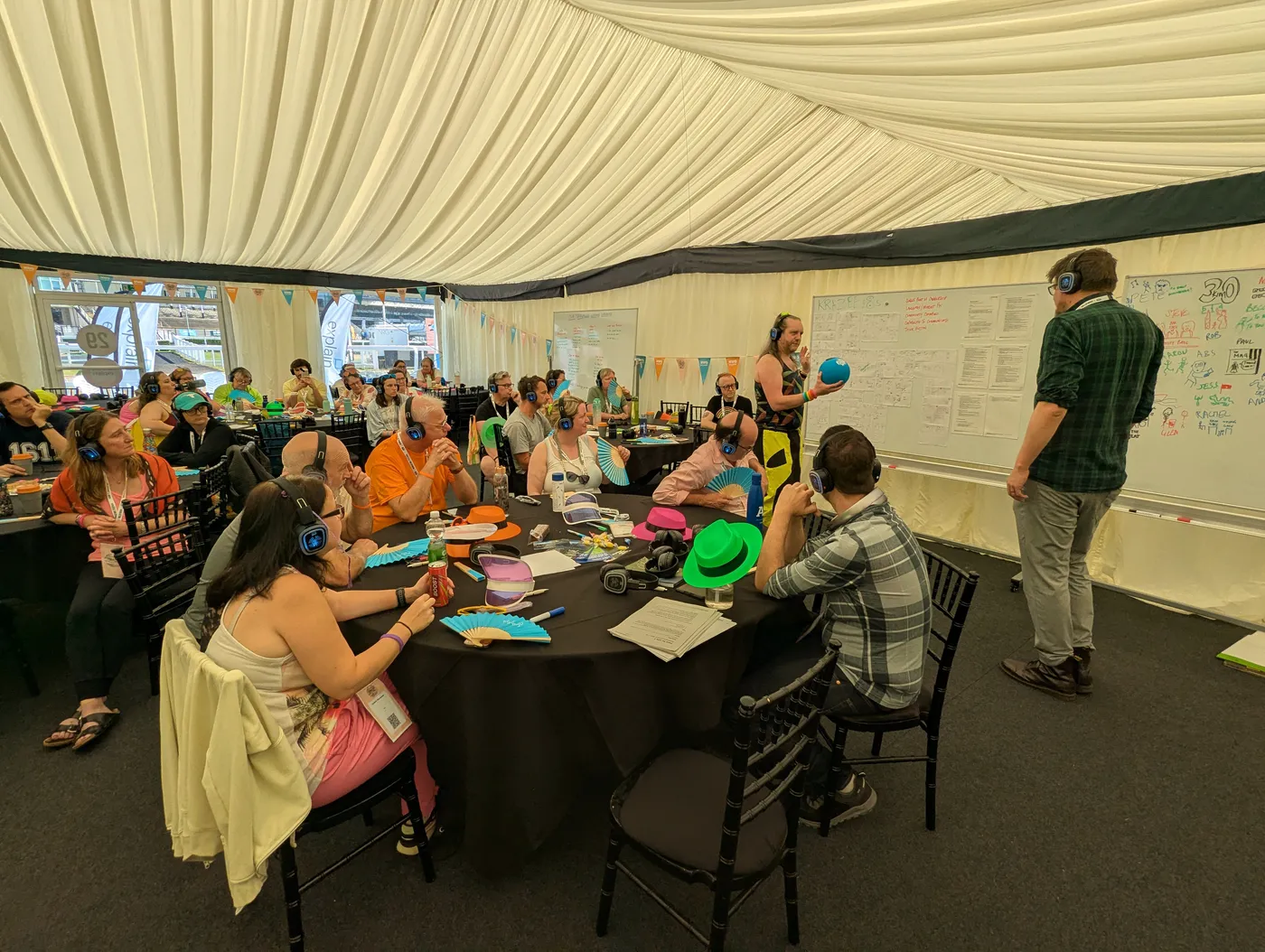 Customers and facilitators working around tables in the Fans for Life 2 sprint room, with whiteboards of sprint output behind them and an Explain flag visible outside