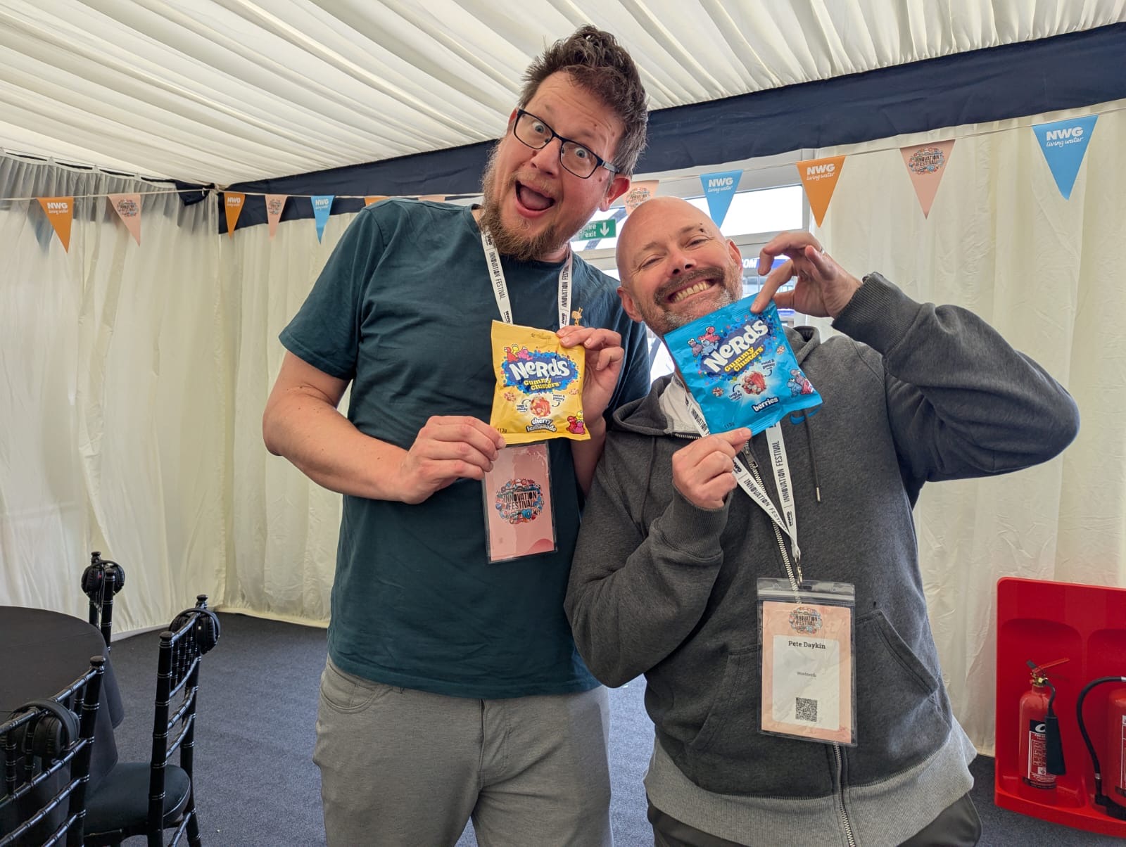Pete Daykin and a colleague grinning in the festival marquee, each holding a packet of Nerds sweets in front of their lanyards
